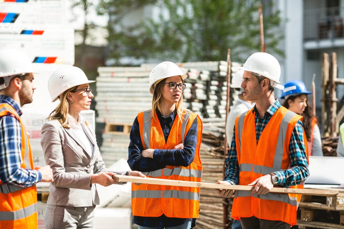 Men and women wearing construction vests and hard hats holding a piece of lumber in a warehouse
