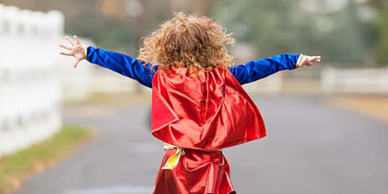 Rear view of a little girl running outside wearing a superhero costume. Her arms are out to each side as she pretends to fly like a superhero.
